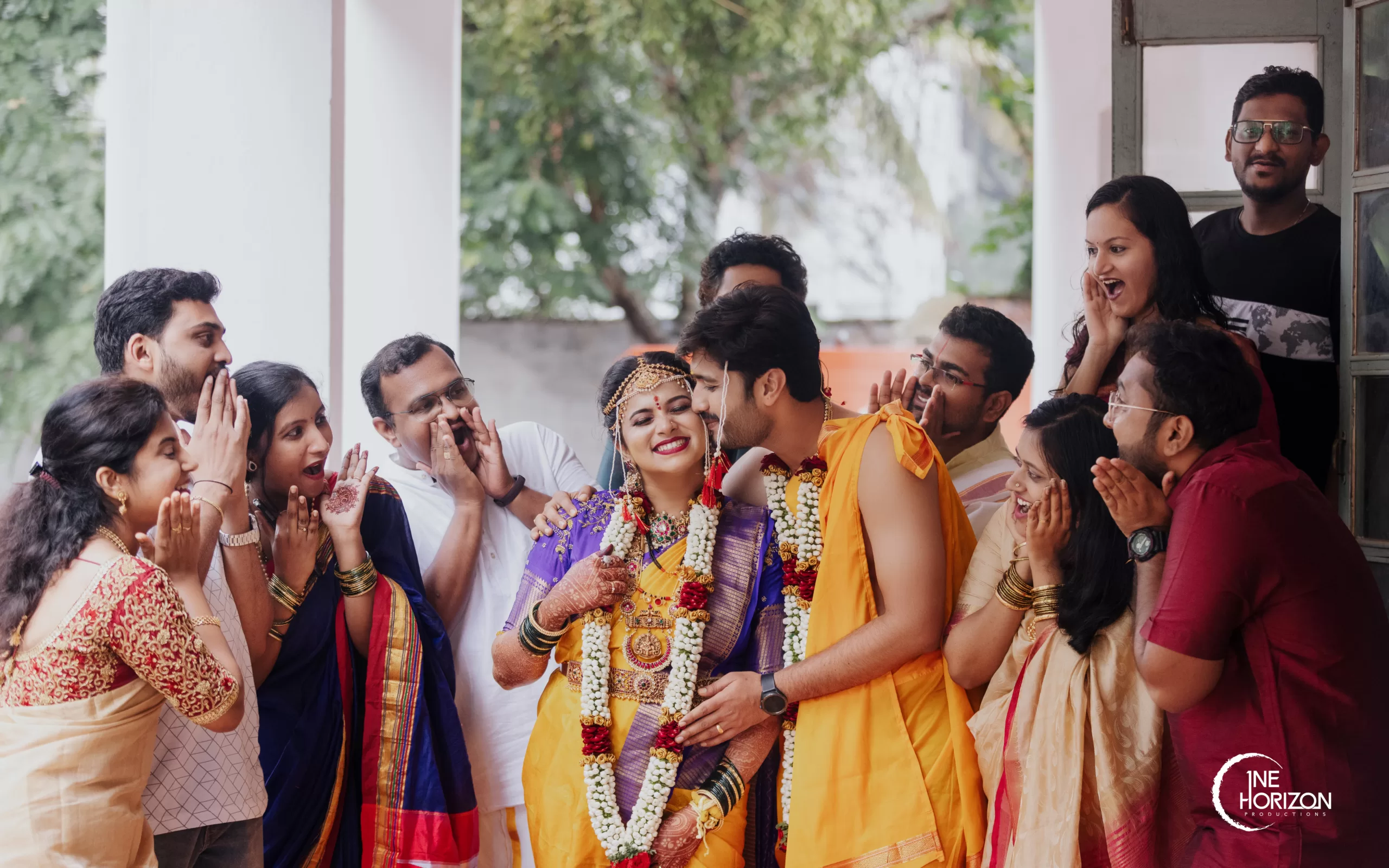 Groom kisses the bride’s cheeks, with family and friends surrounding them with fun expressions.